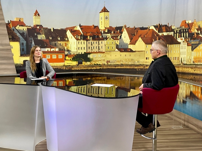 Eine Person sitzt am Studiotisch vor dem beleuchteten Hintergrund. Die Atmosph&auml;re wirkt professionell und ruhig.