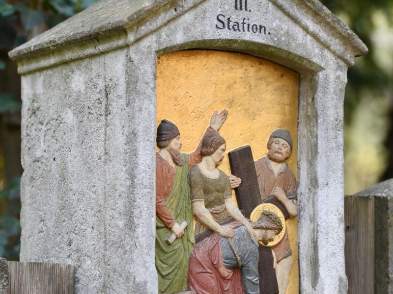 Steinkapelle mit Kreuz auf dem Dach, die die siebte Station des Kreuzwegs darstellt. Die Nische zeigt Jesus, der unter dem Kreuz zusammenbricht, umgeben von Soldaten und weinenden Frauen. Die Inschrift lautet "Jesu f&auml;llt das 2te mal unter dem Kreuze.