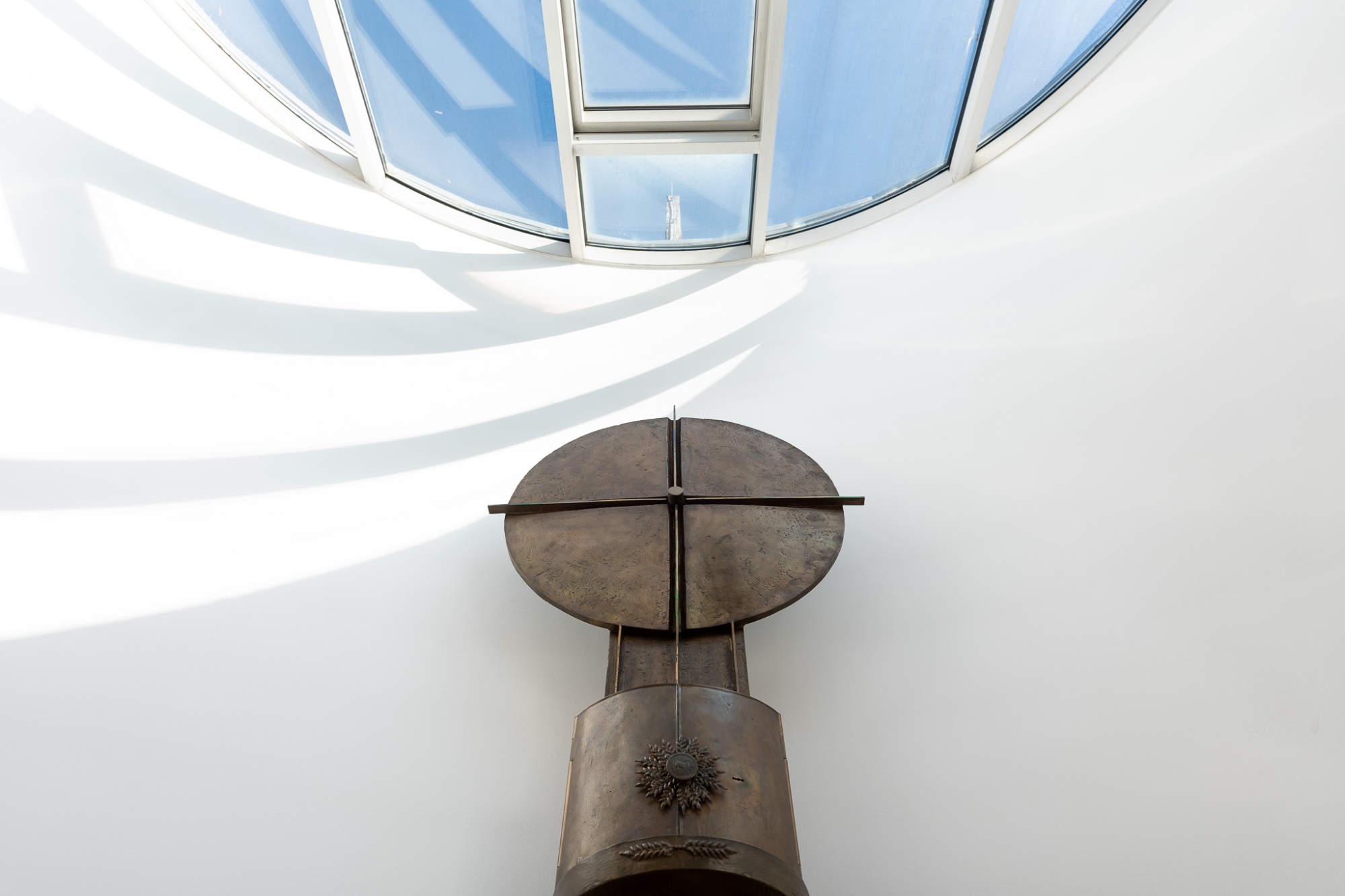 Ein Blick von unten auf einen modernen Kronleuchter mit geometrischen Metallelementen und Glaseinsätzen, durch die blauer Himmel sichtbar ist, vor einer weißen Decke mit geschwungenen Architekturlinien.