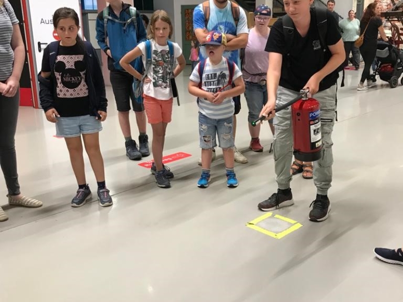 An einer Ausstellung stehen Besucher verschiedenen Alters um einen &auml;lteren Mann herum, der technische Ger&auml;te auf einem Tisch pr&auml;sentiert. Kinder und Erwachsene beobachten interessiert die Demonstration in einem Museum oder Ausstellungsraum mit roten und blauen W&auml;nden.